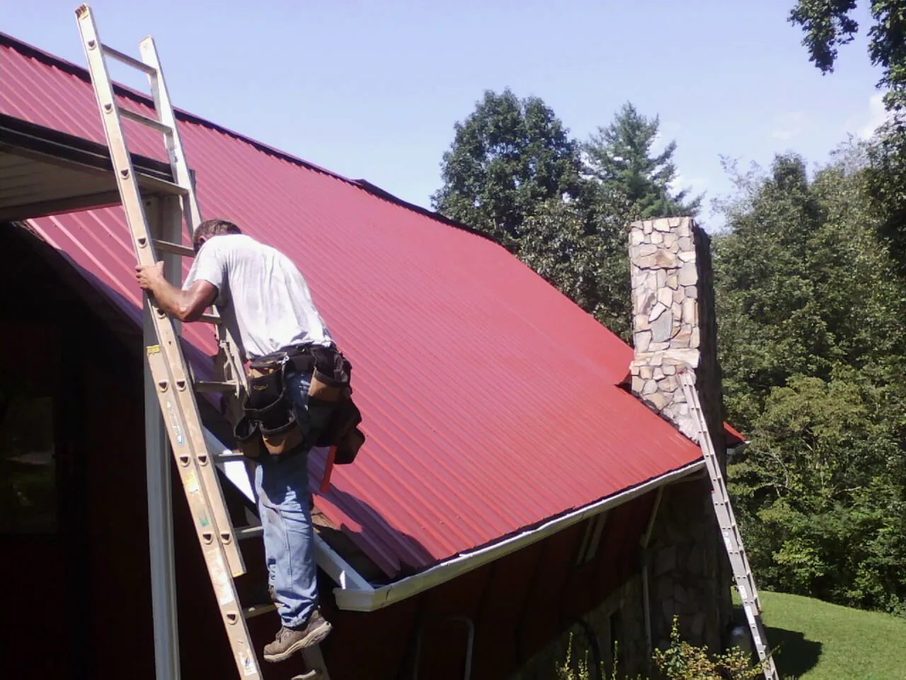 Roofer on a ladder beside a red metal roof and stone chimney — Soffit & Fascia Repair by Roofing Brothers in Honeyville